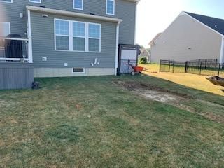 Backyard with house, deck, and green lawn. Wheelbarrow near side door, small black fence in the distance.