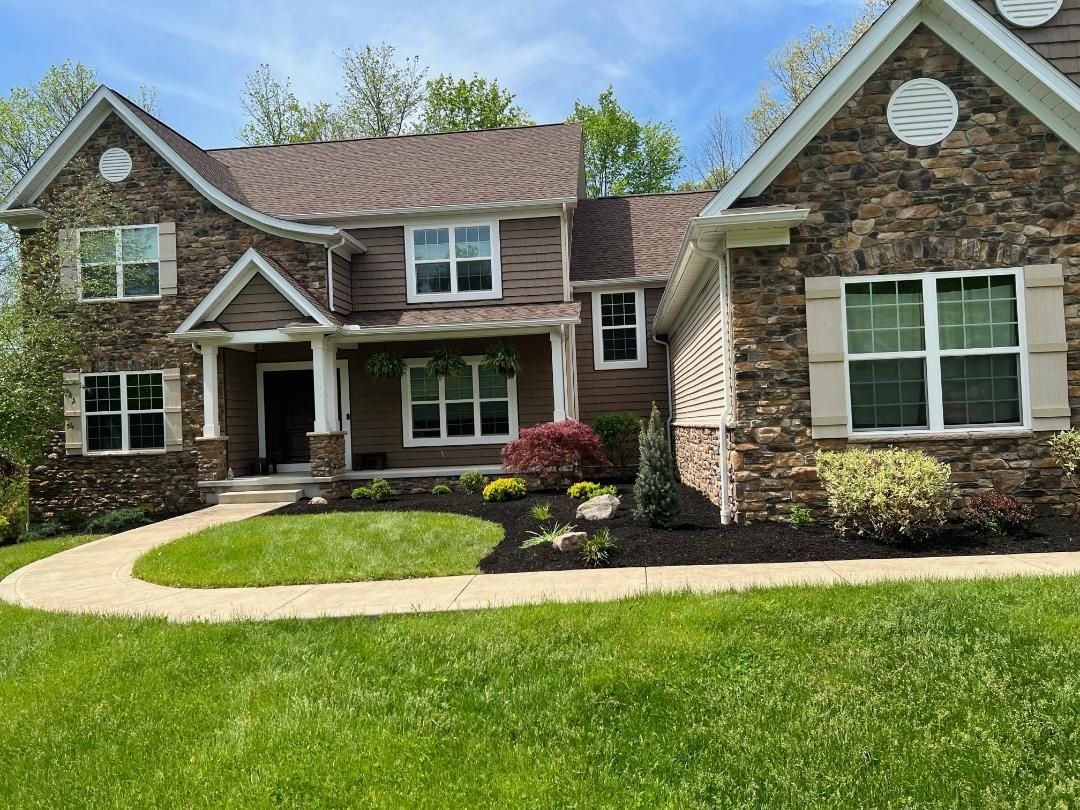 Two-story house with brown and stone exterior, manicured lawn, and landscaping under a blue sky.