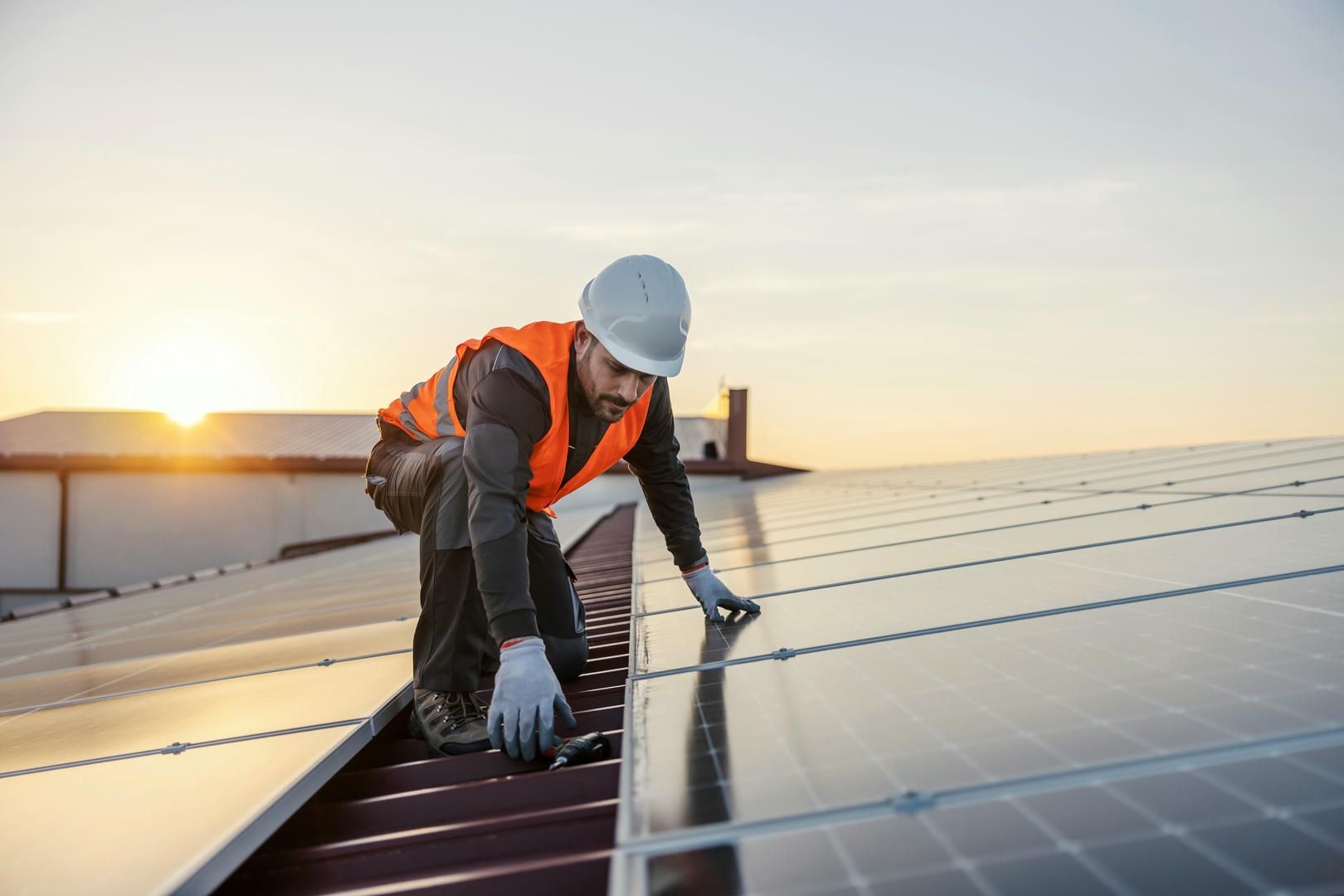 Rooftop solar panel installer in orange vest and hard hat working on panels during sunset.