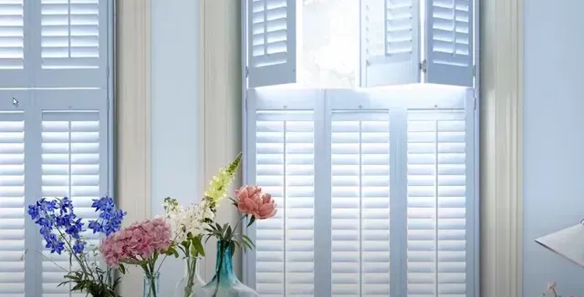 A vase of flowers sits on a table in front of a window with white shutters. Love is Blinds Arizona (844) 936-2446