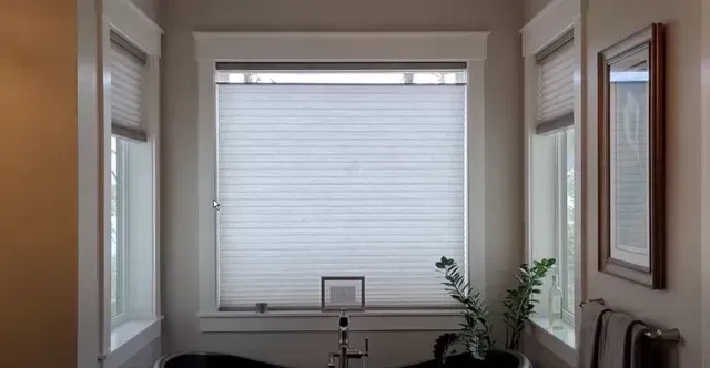 A bathroom with a tub and a window with honeycomb shades.