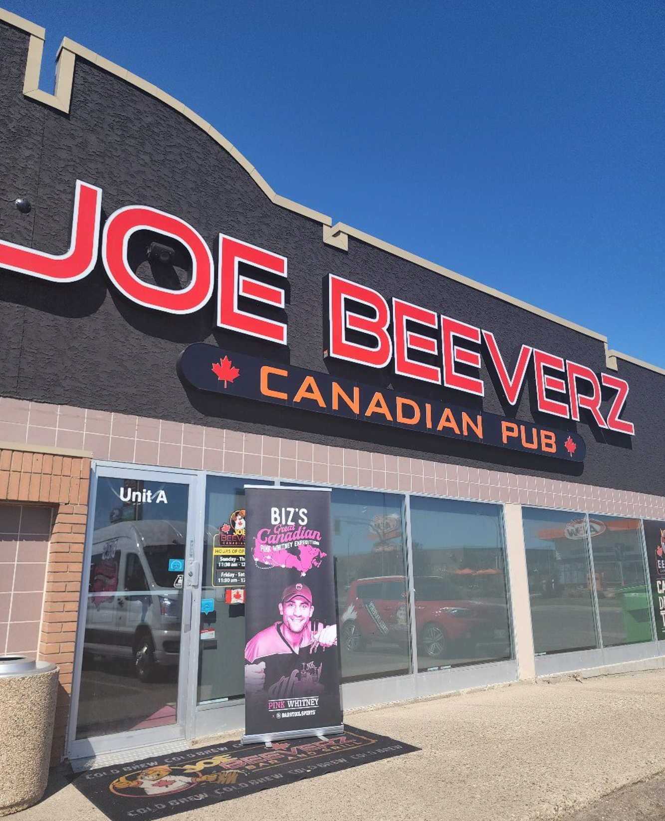 Joe Beeverz Canadian Pub storefront with red neon sign and a sidewalk poster display under a blue sky
