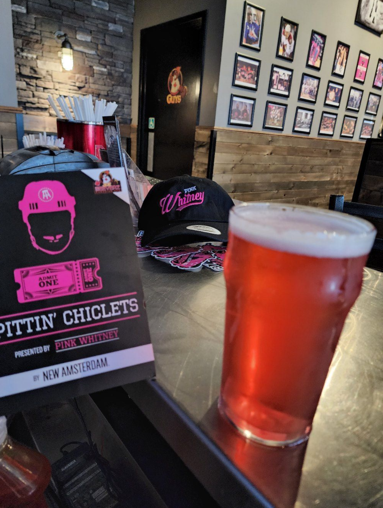 Pint glass of amber beer on a table beside a restaurant display and black cap with pink lettering