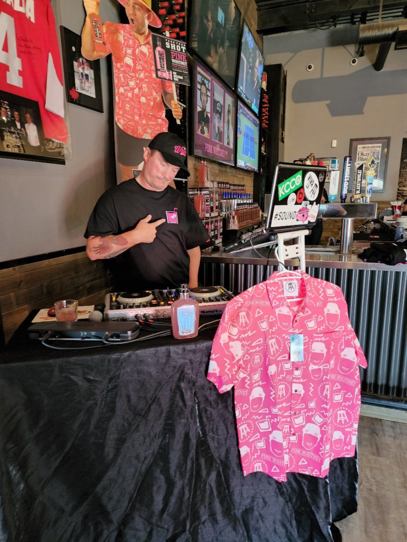 Vendor behind a black table at a market stall with pink shirts hanging in front.
