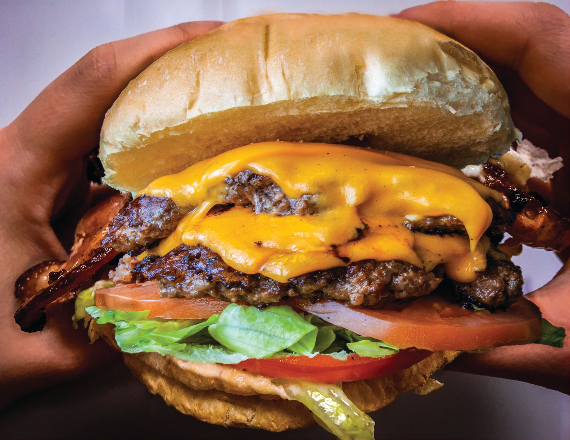 Hands holding a cheeseburger with melted cheese, bacon, lettuce, and tomato on a bun