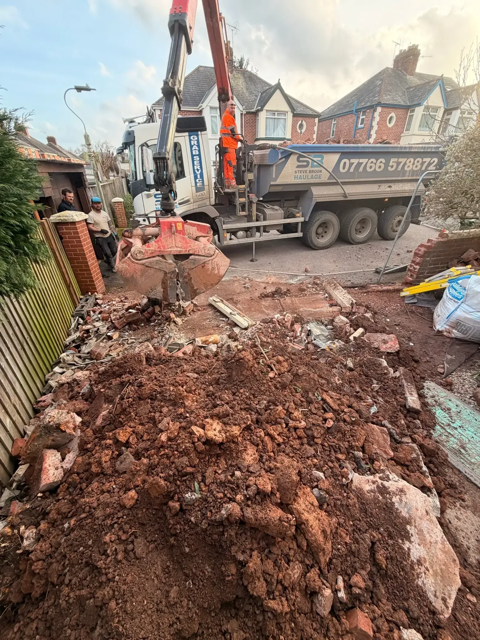 Roadworks with excavator and truck on a muddy residential street, rubble piled in foreground