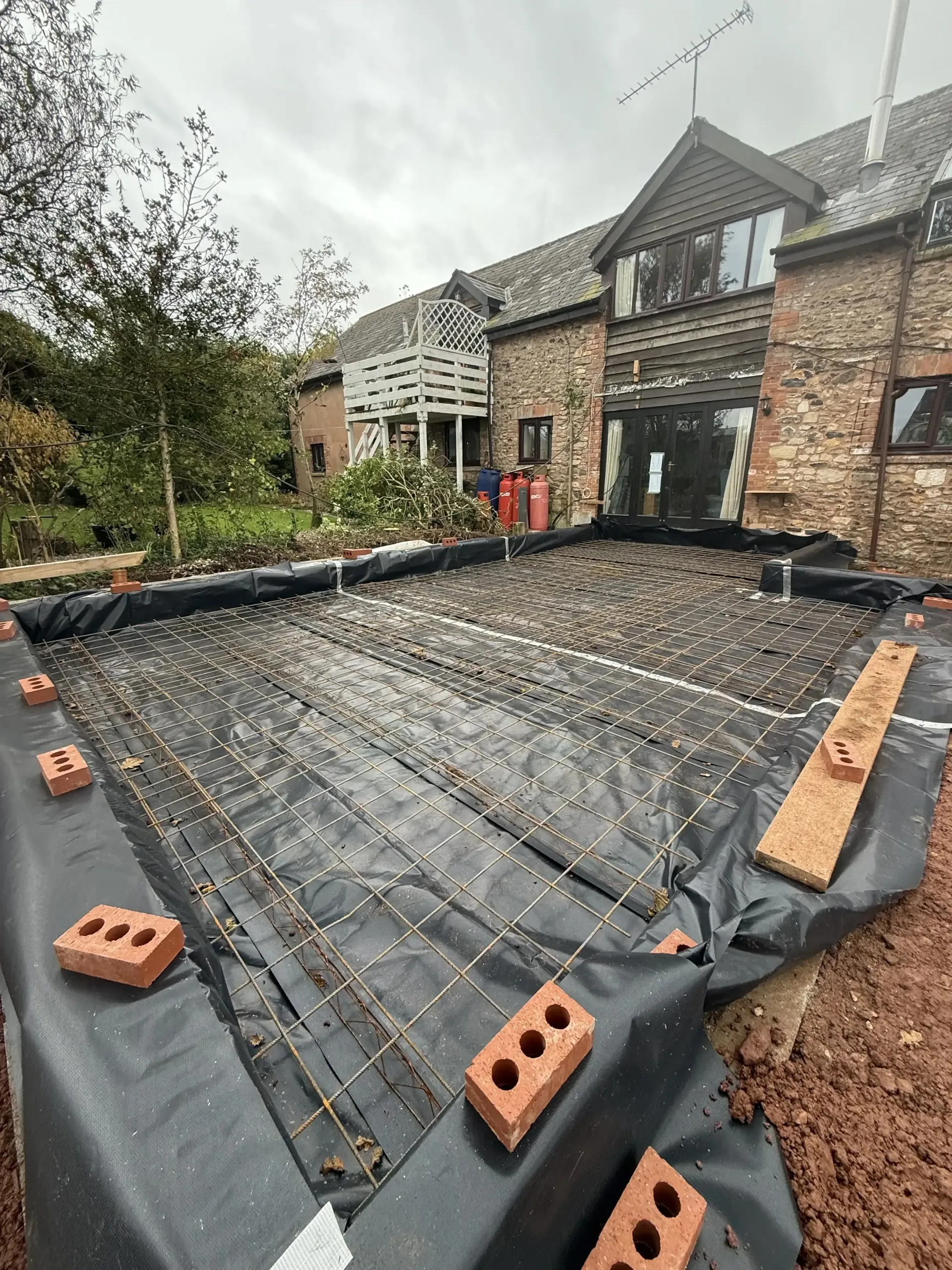 Reinforced foundation slab under construction beside a brick house, with rebar, black sheeting, and bricks around the edges