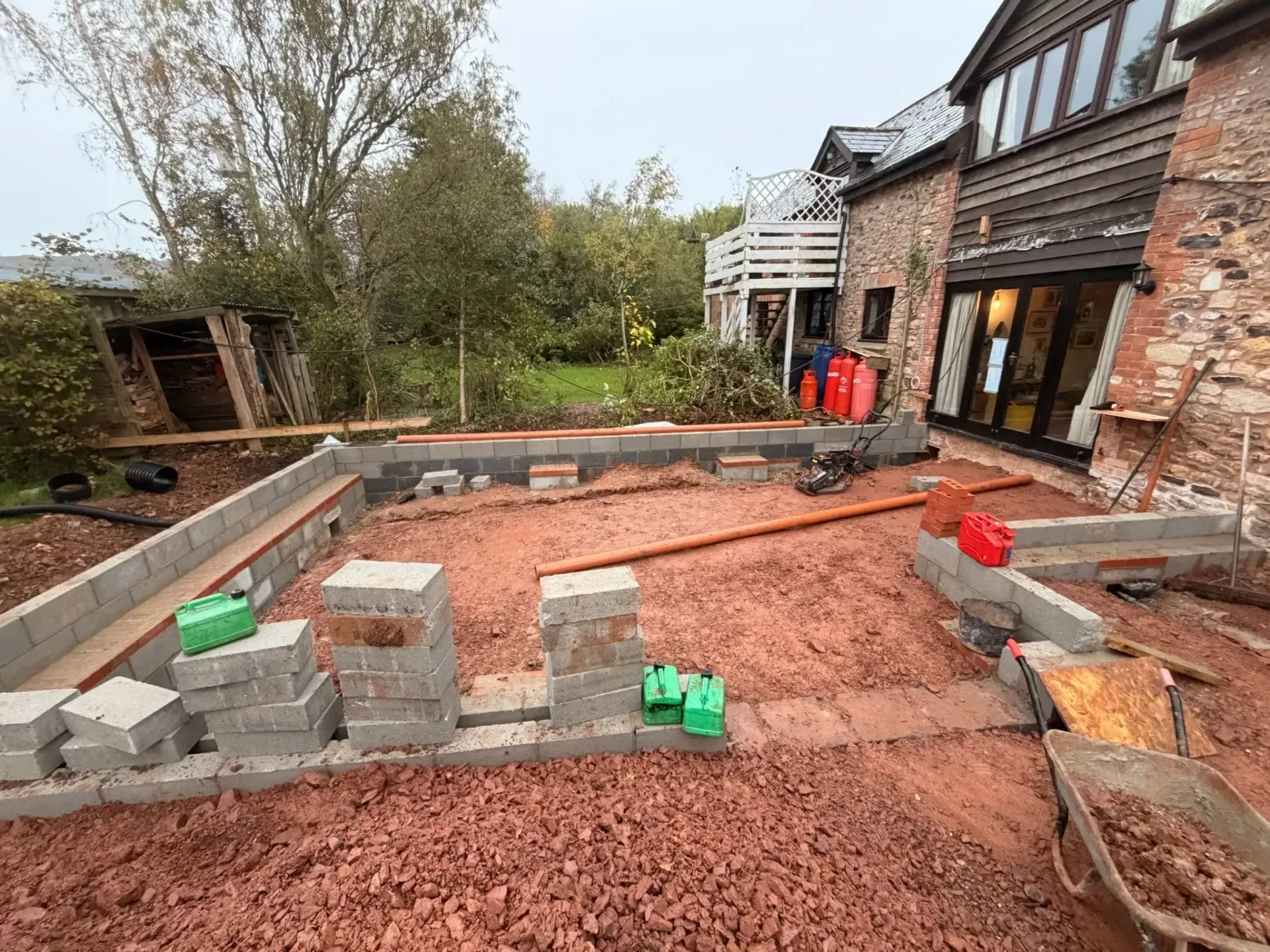 Backyard patio under construction beside a brick house, with red gravel, concrete blocks, and tools.