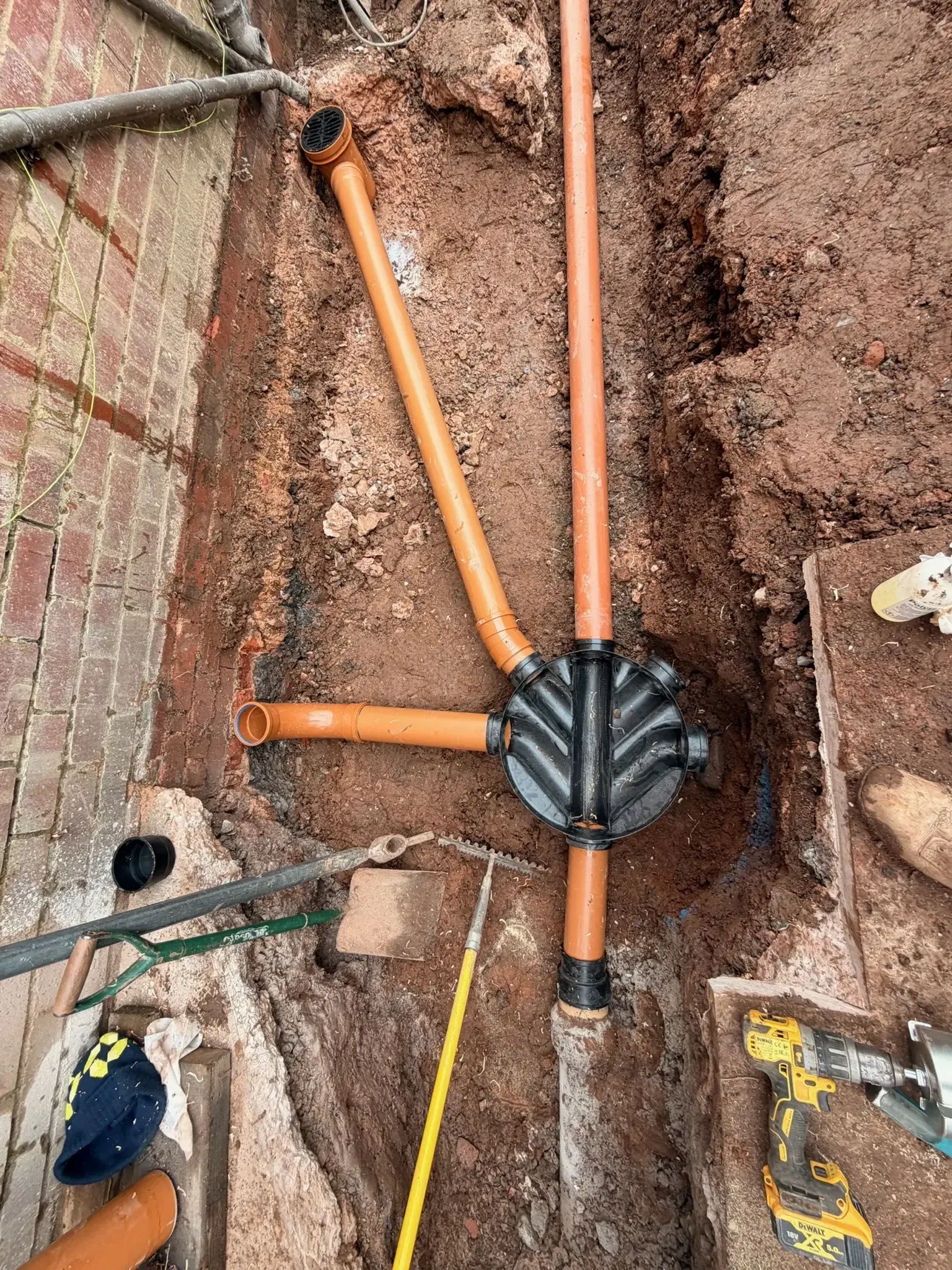 Orange plumbing pipes and a black junction fitting installed in a dirt excavation trench