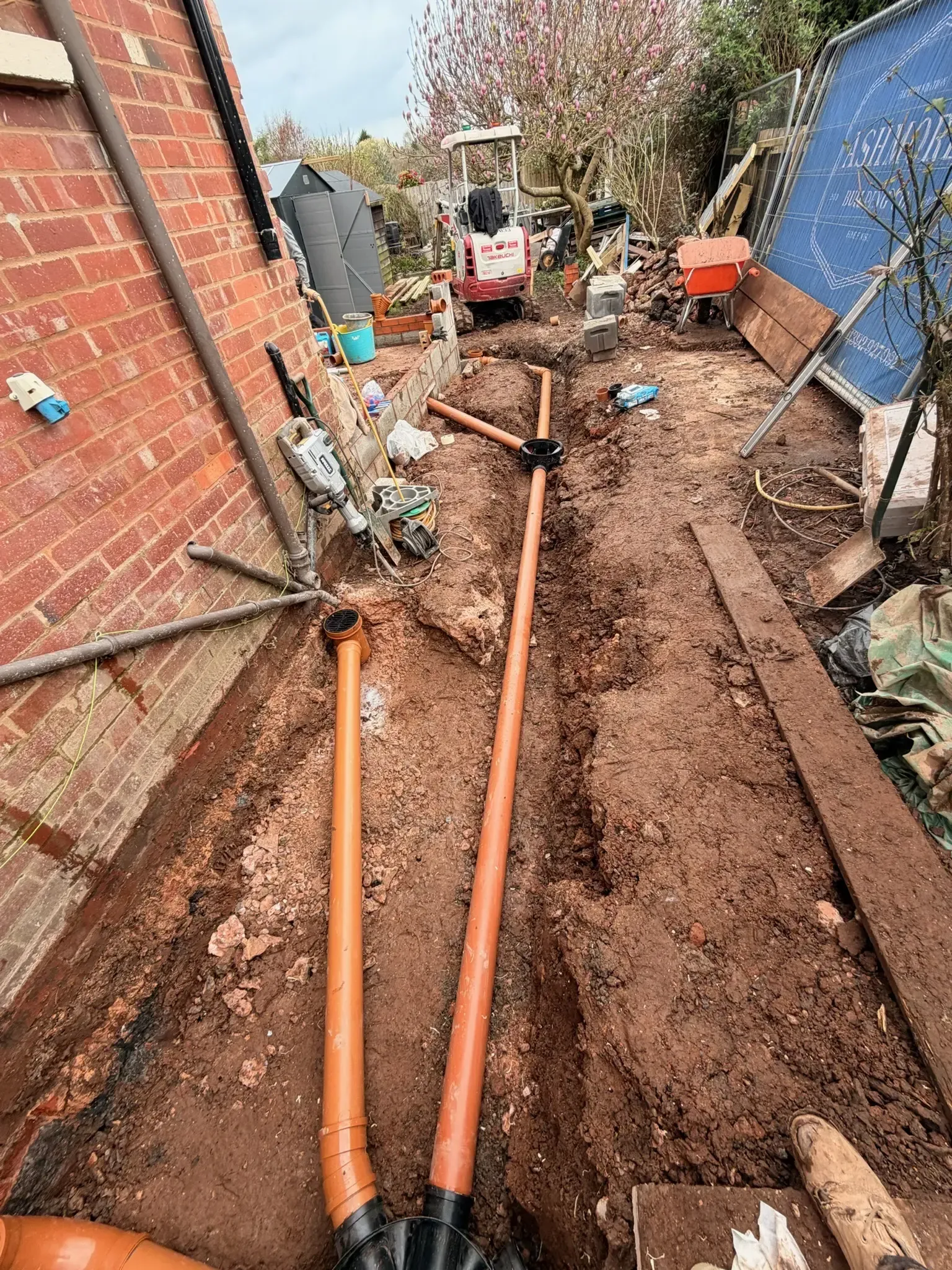 Backyard trench with two orange pipes laid in muddy ground beside a brick house.