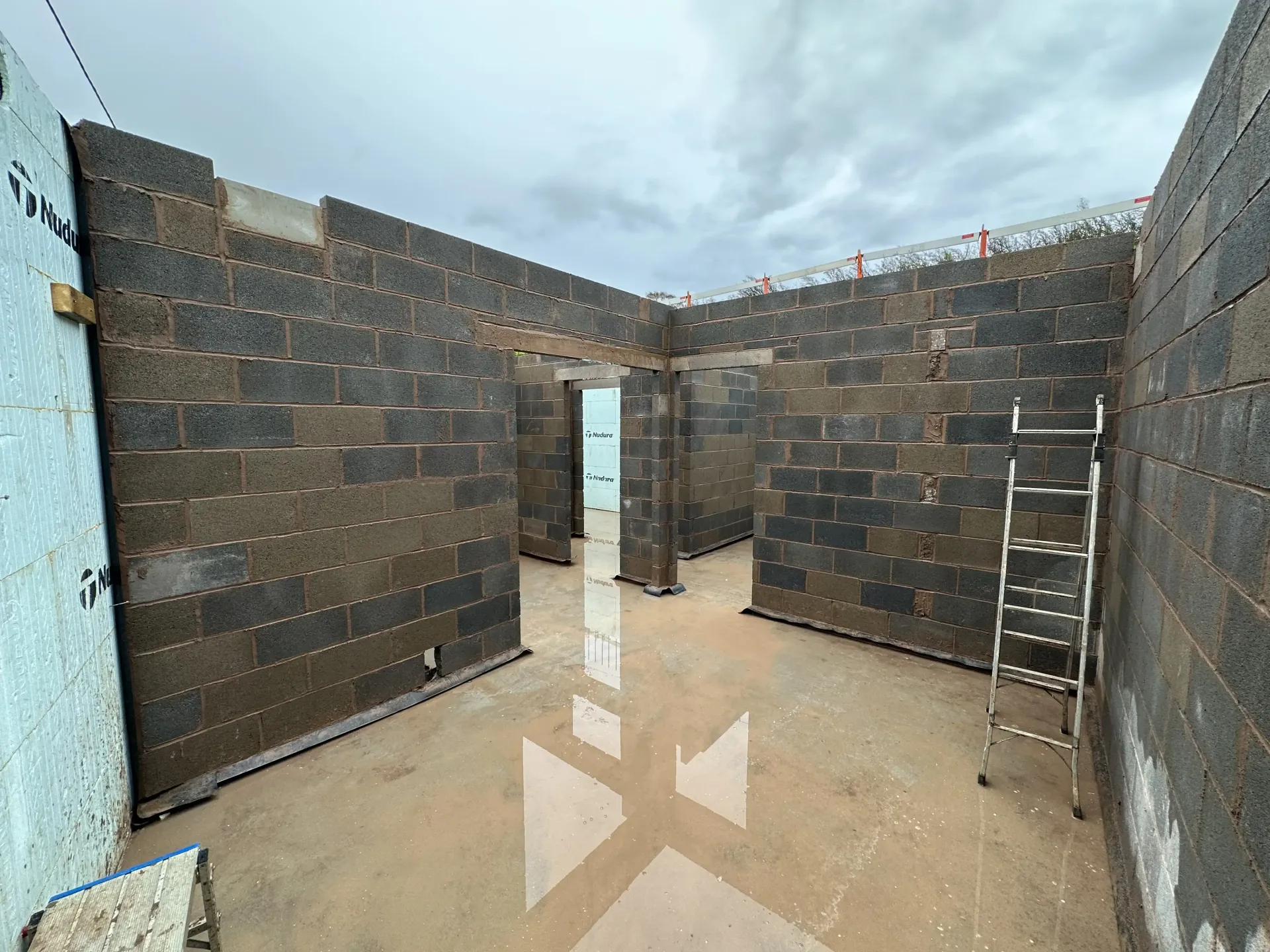 Partially built room with brick walls, open doorway, and ladder on a wet concrete floor
