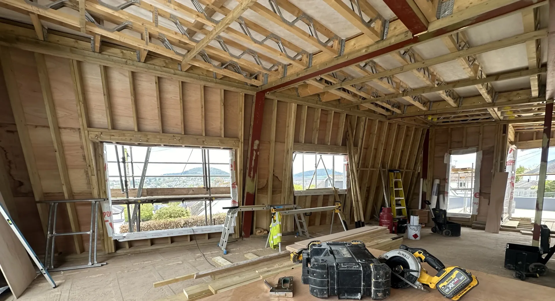 Interior of a house under construction with exposed framing, large window openings, and scattered tools and materials.