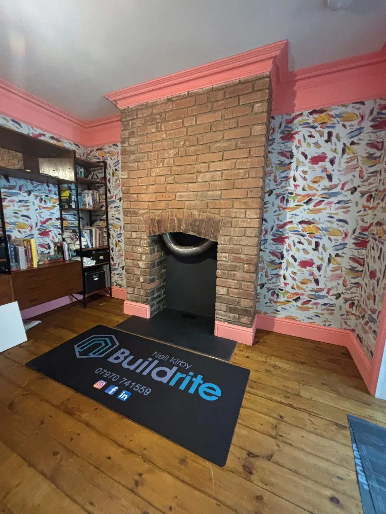 Brick fireplace in a pink room with patterned wallpaper, wooden floor, and a black Buildsite mat.