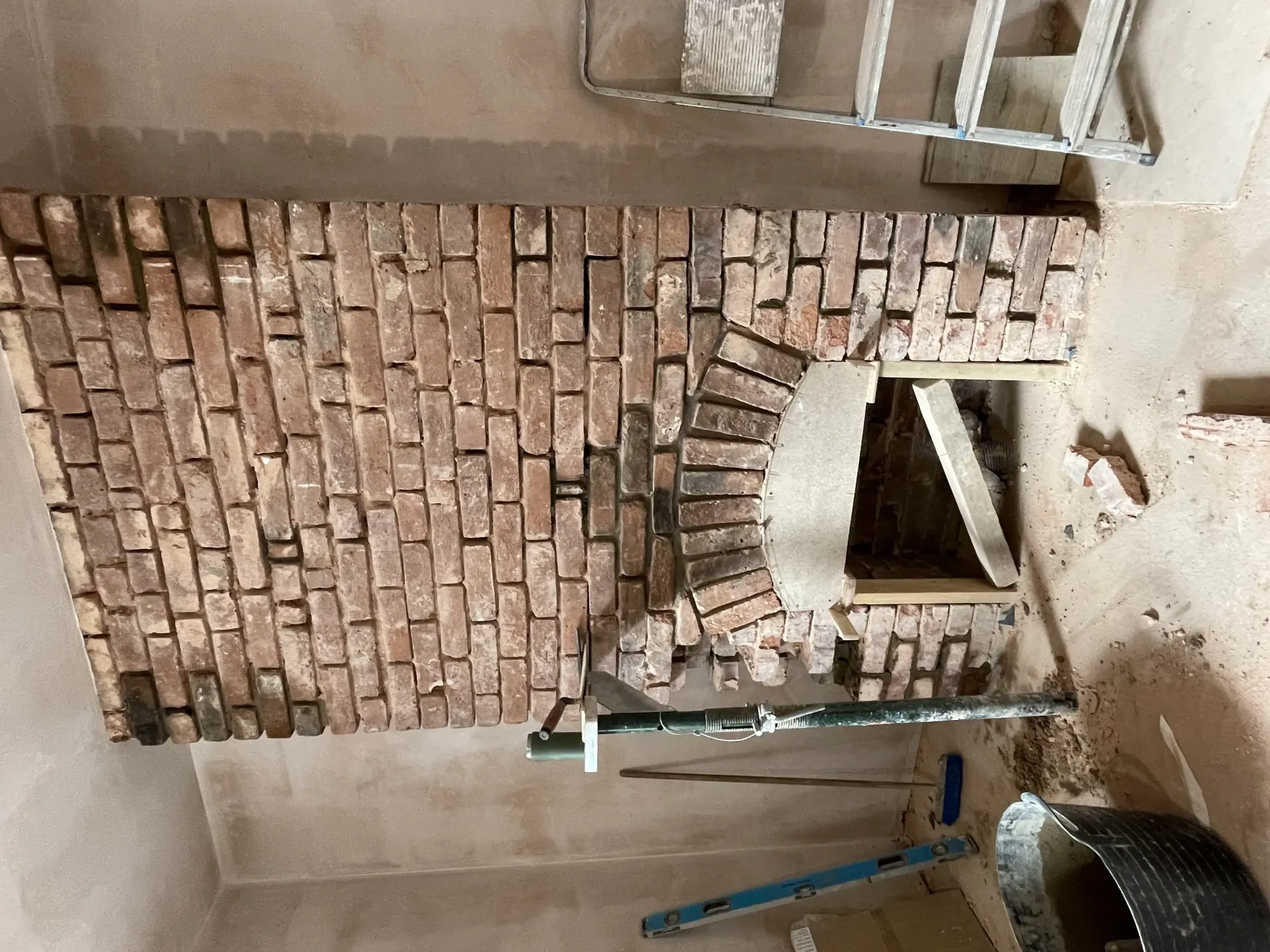 Partial brick wall under construction with a circular opening, ladder, and cement floor nearby