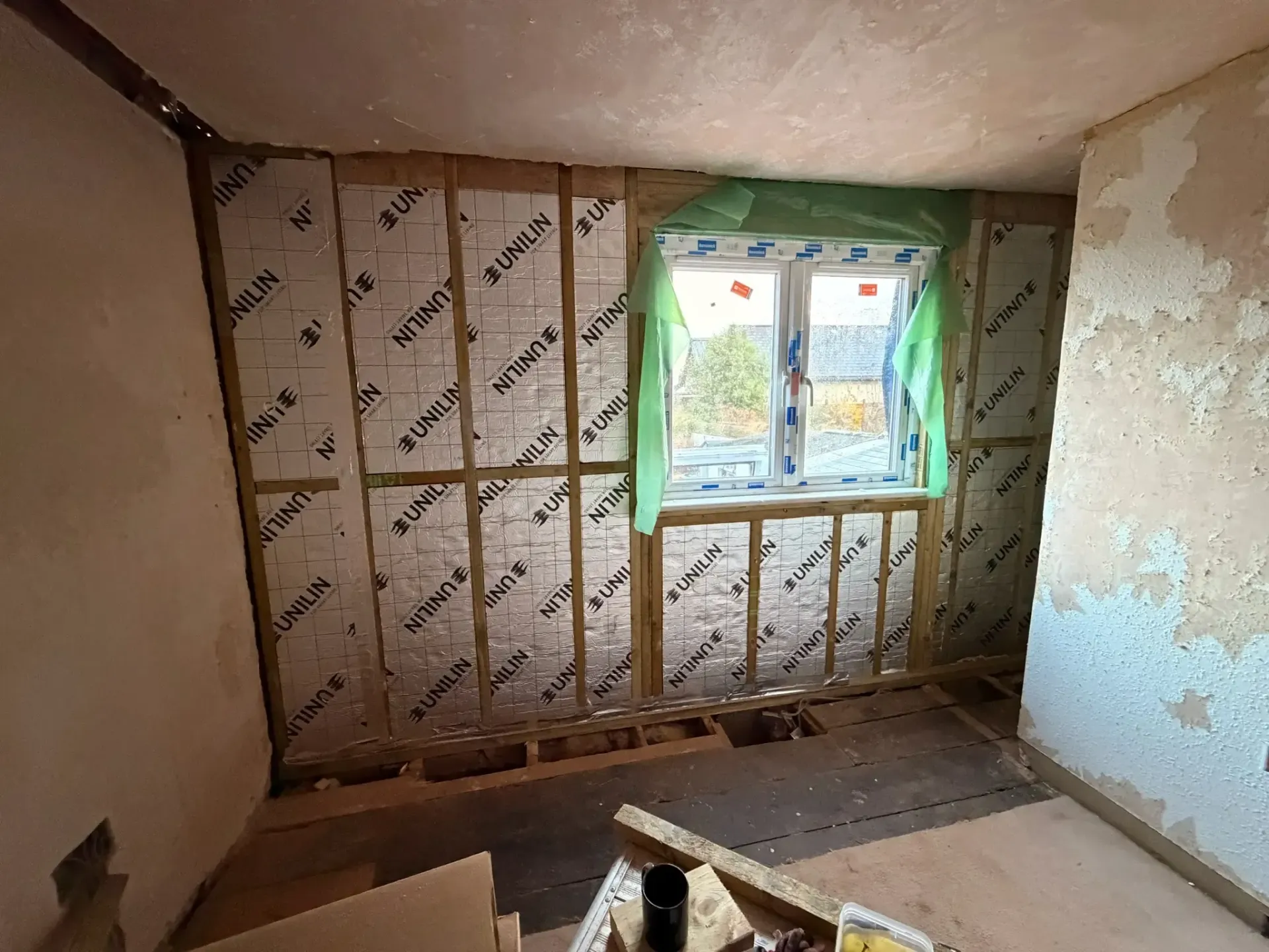 Partially stripped room with exposed wall studs, peeling plaster, and a window letting in daylight.
