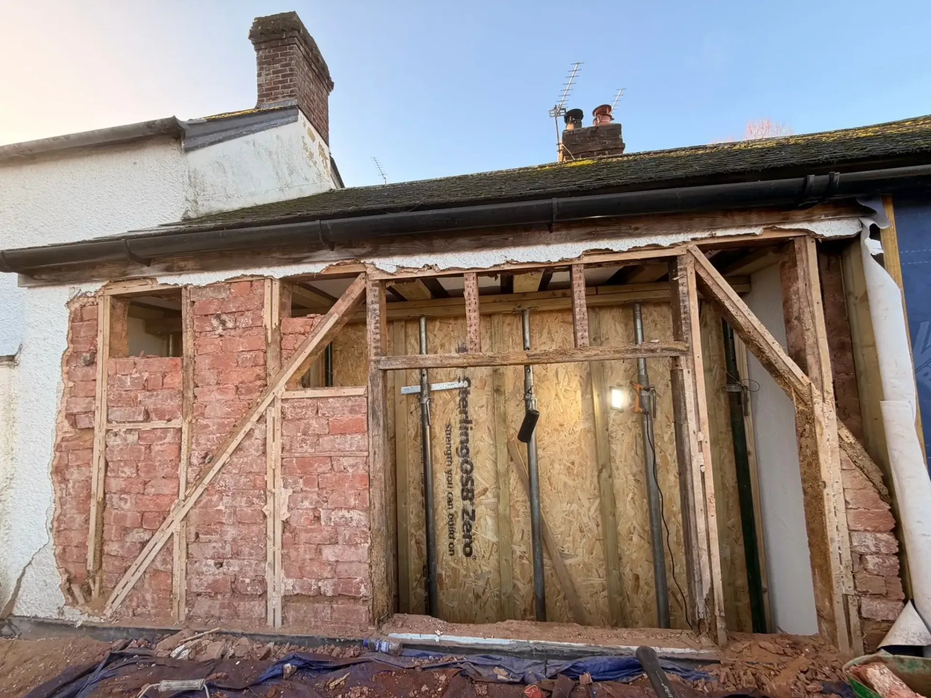 Partially demolished house wall with exposed pink insulation and wooden framing under a tiled roof