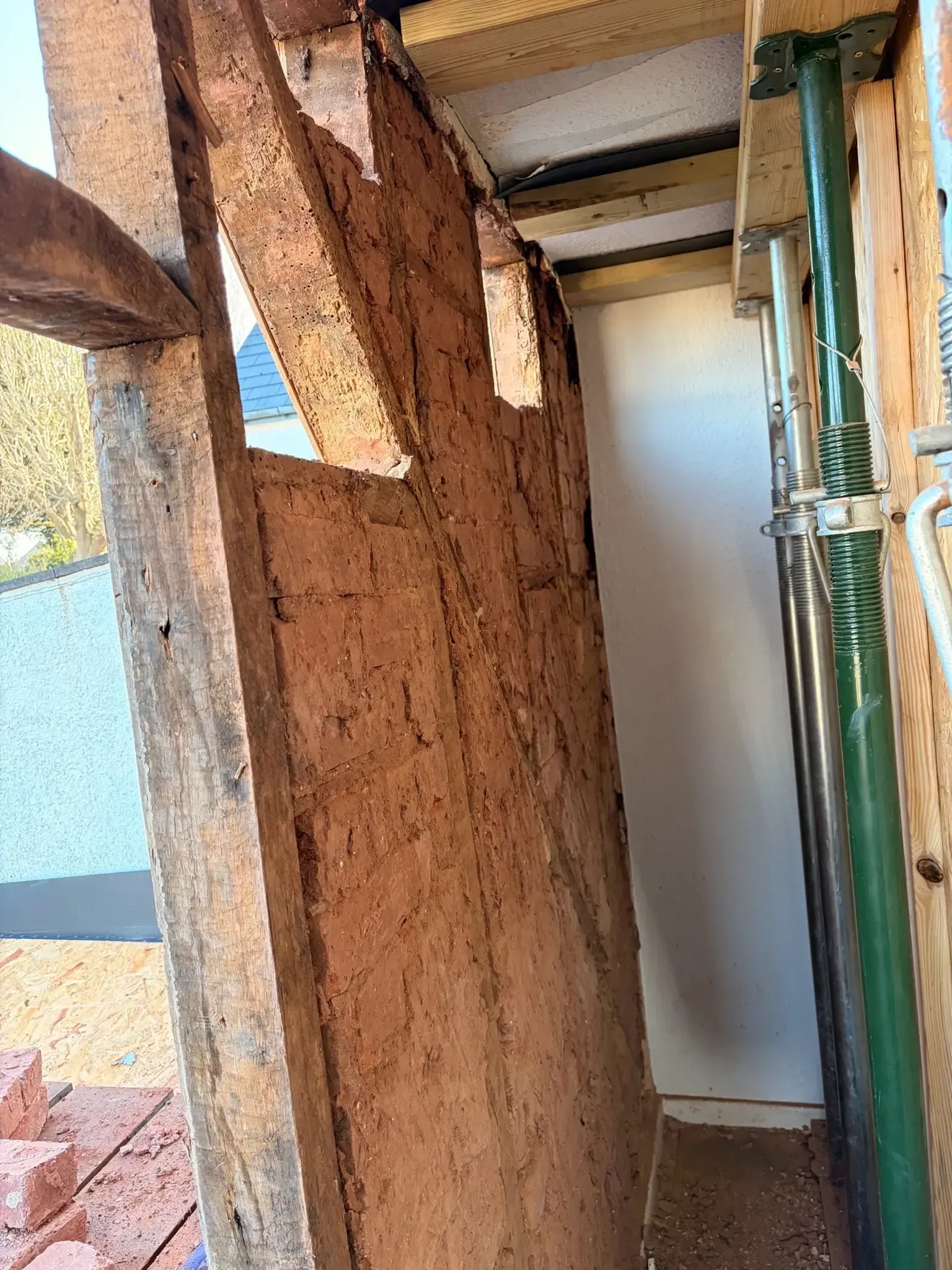 Damaged mud wall in a narrow outdoor corridor, with exposed wood framing and a white door in the background