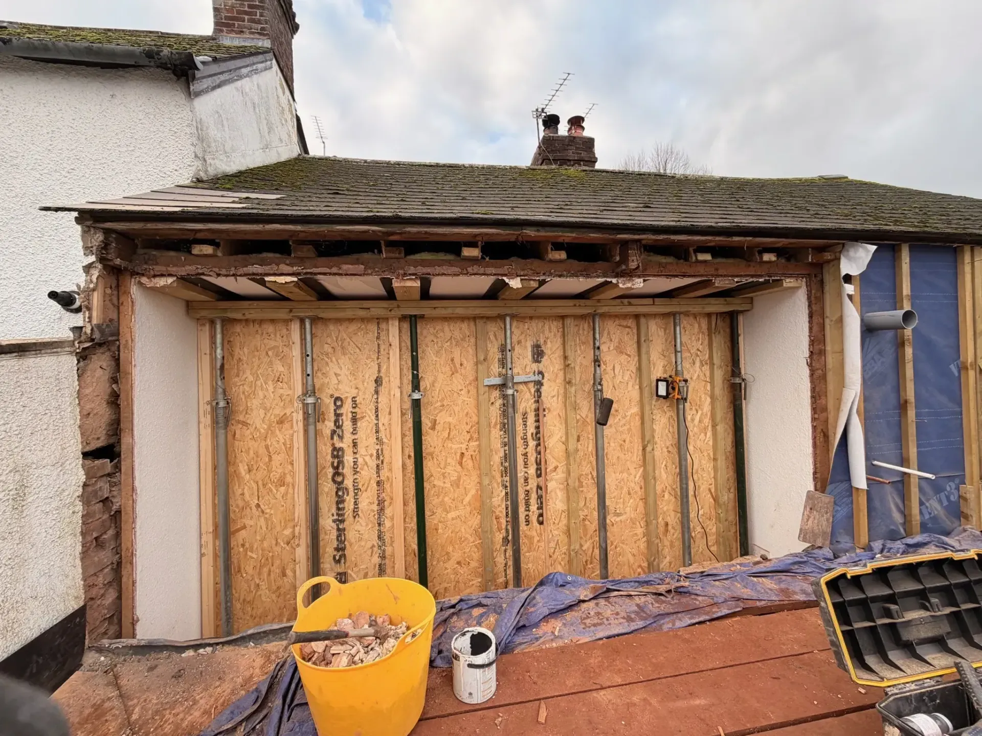 Damaged garage with exposed wooden framing, debris, and a yellow bucket in front