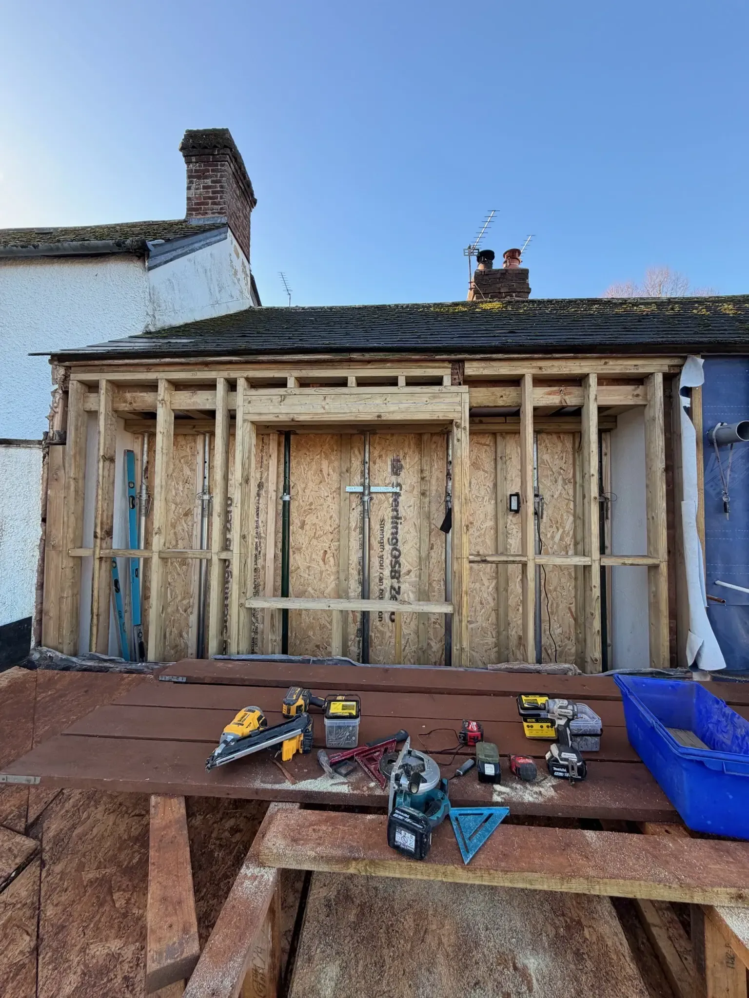House exterior under renovation with exposed framing, tools on a deck, and a blue tarp on the right