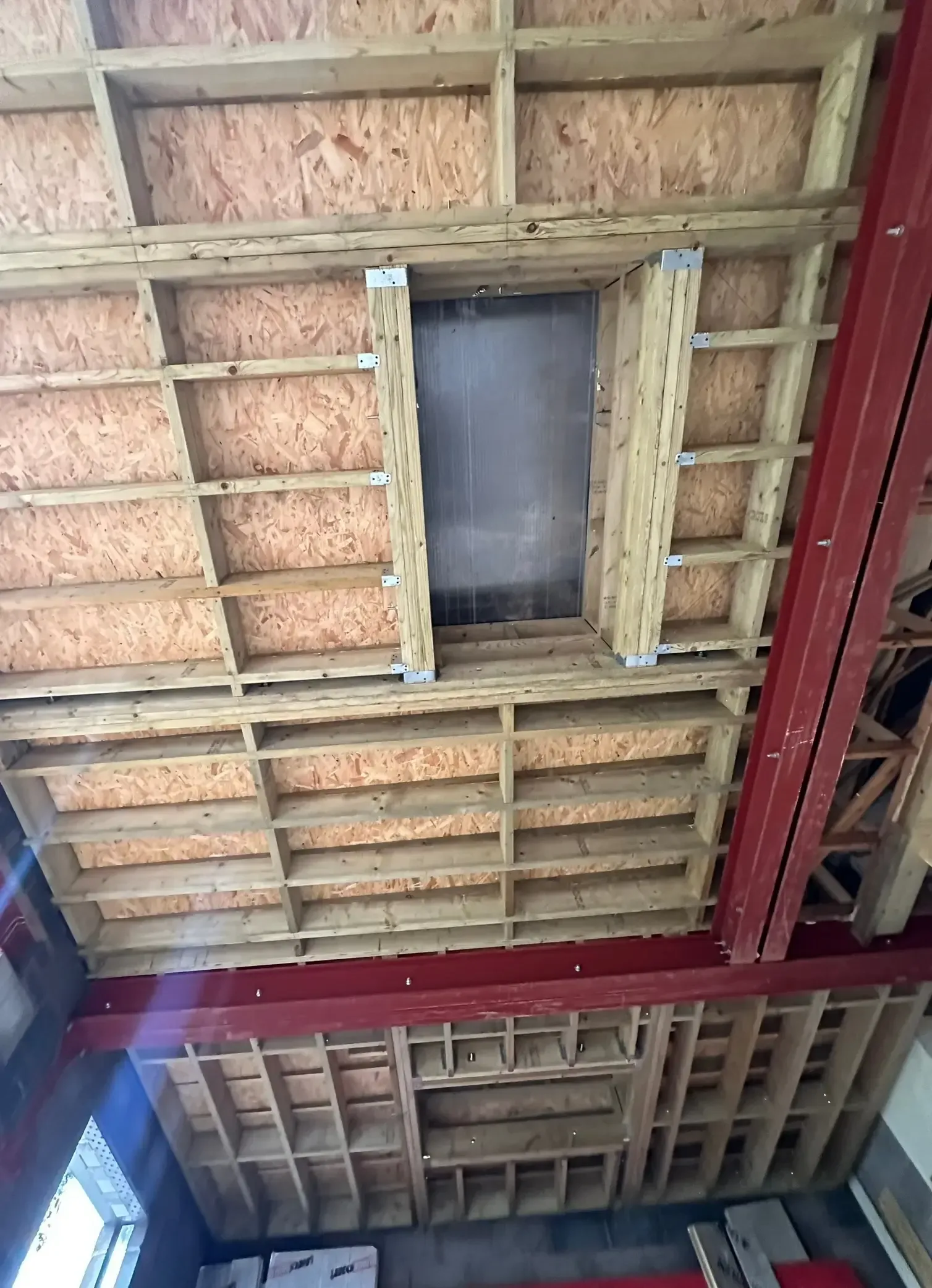 Exposed wooden ceiling framing with insulation and a small rectangular opening above a red steel beam.