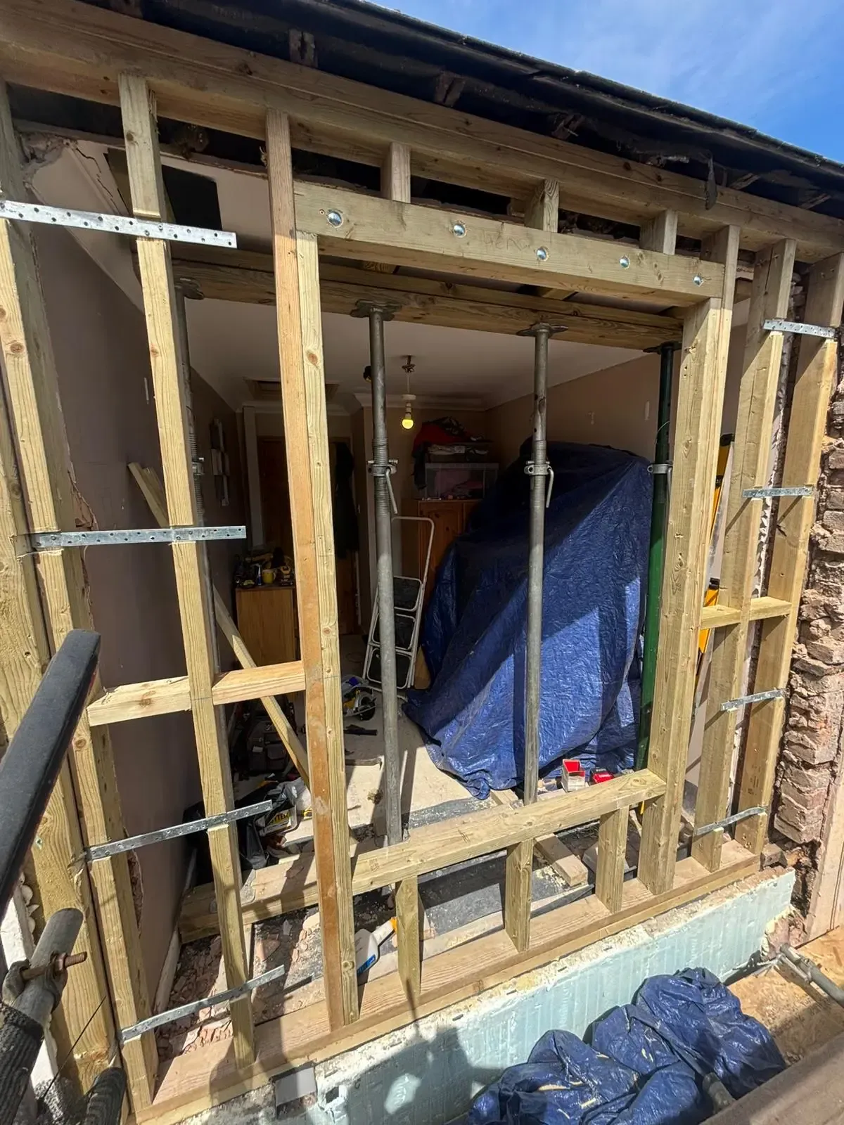 Partially demolished wall with exposed wooden framing and a blue tarp-covered object inside