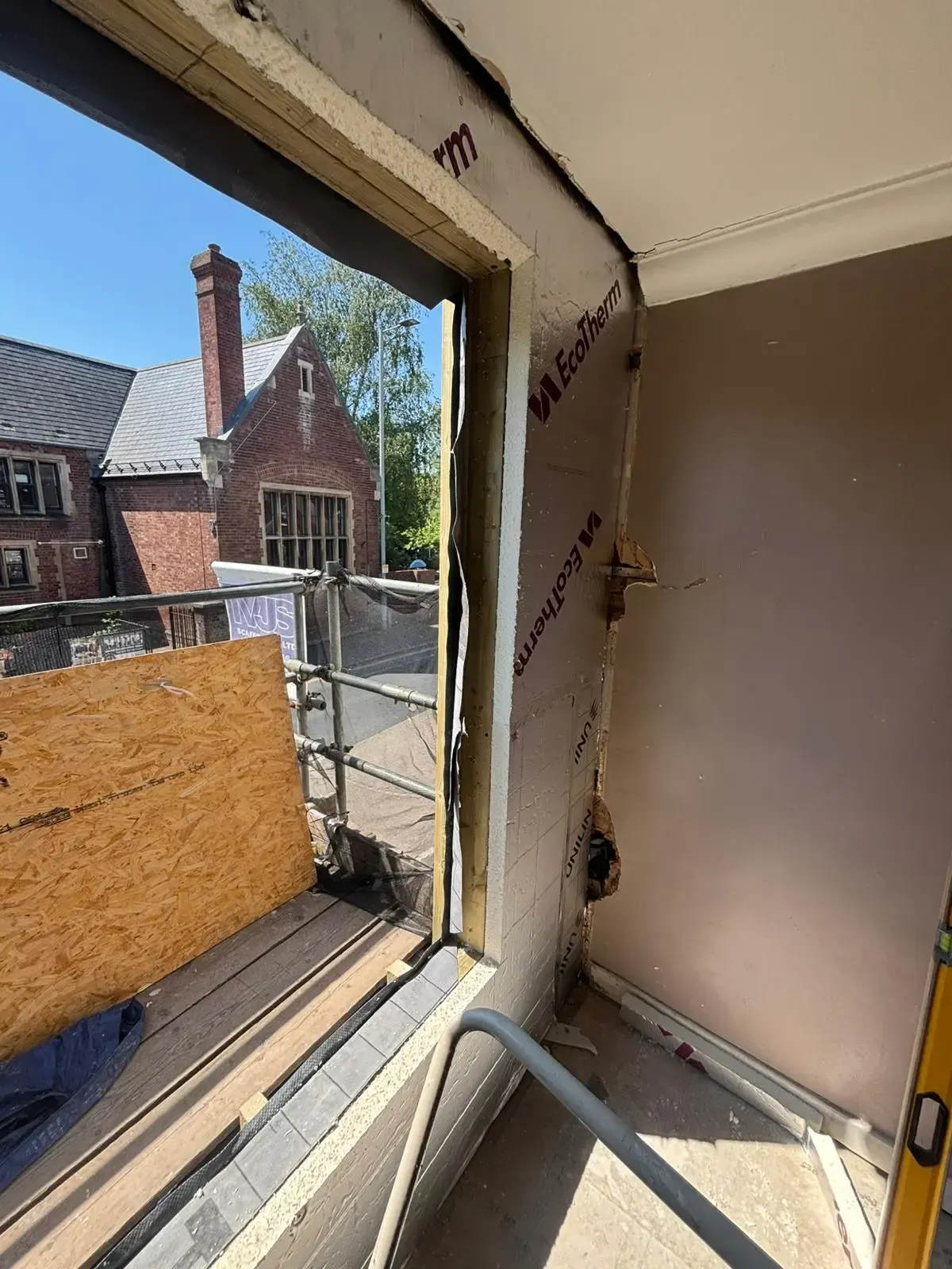 Damaged interior wall beside a window opening, with exposed framing and outdoor view