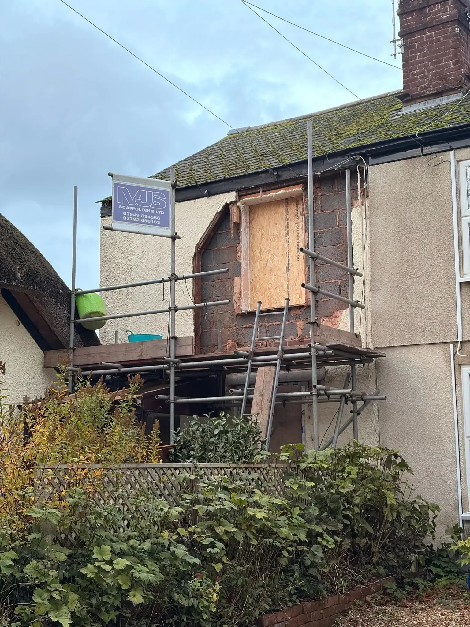 Scaffolded stone house with boarded upper window, hedge in front, and a blue sign on the wall.