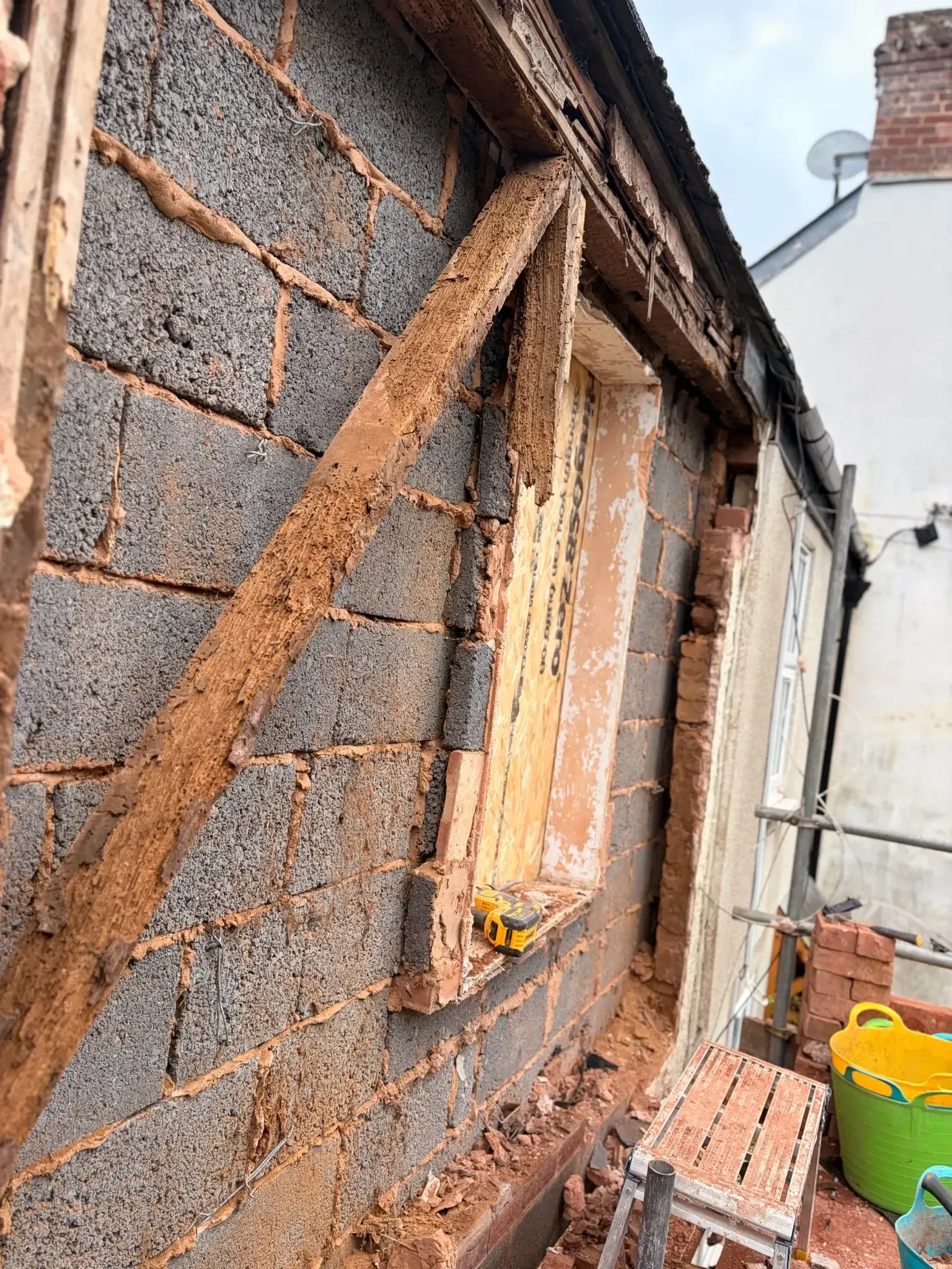 Damaged exterior wall with exposed wooden framing and partially boarded window, next to buckets and tools