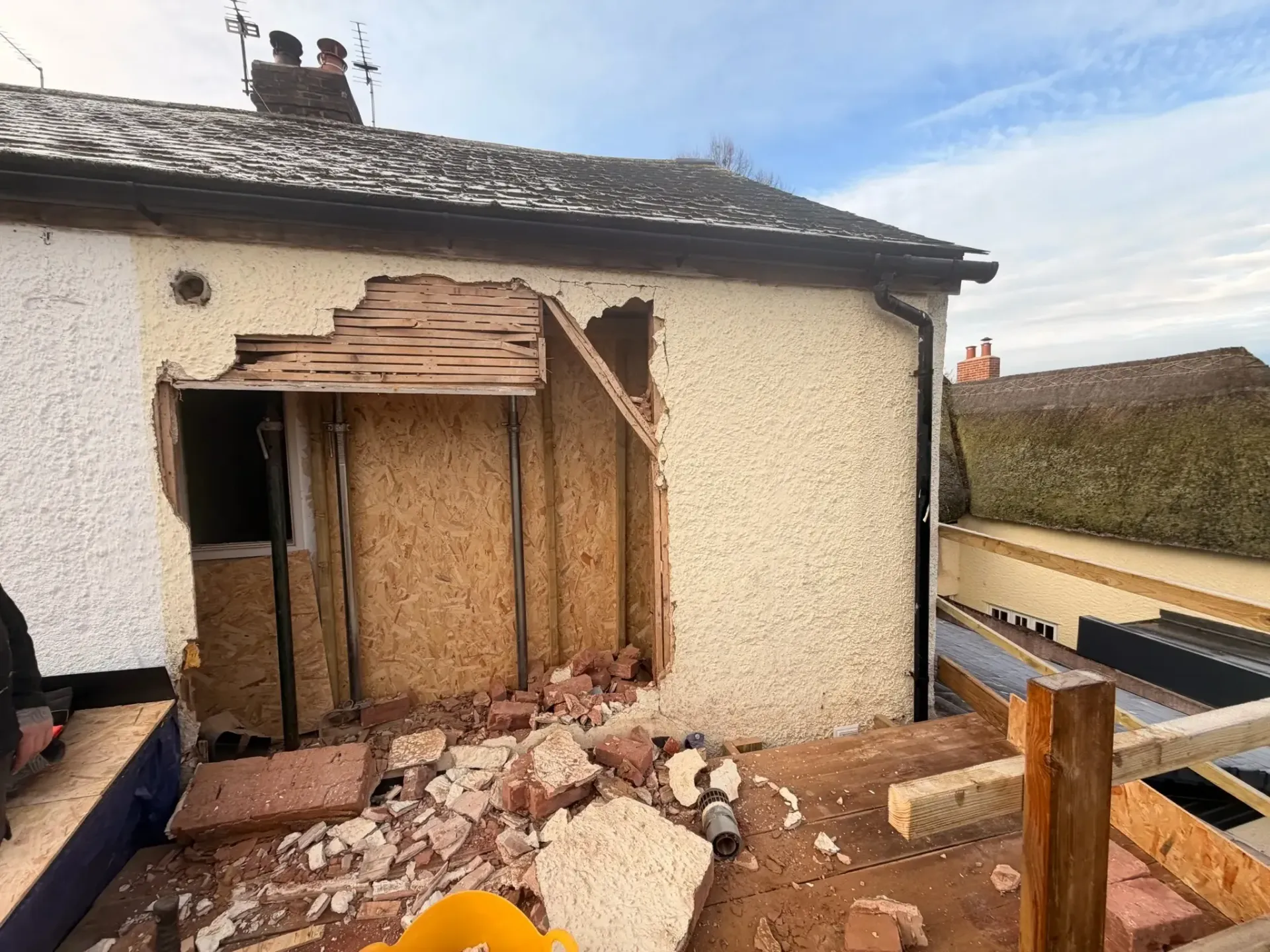 Roof damage and rubble at a house opening, with exposed wooden framing and broken brick debris.