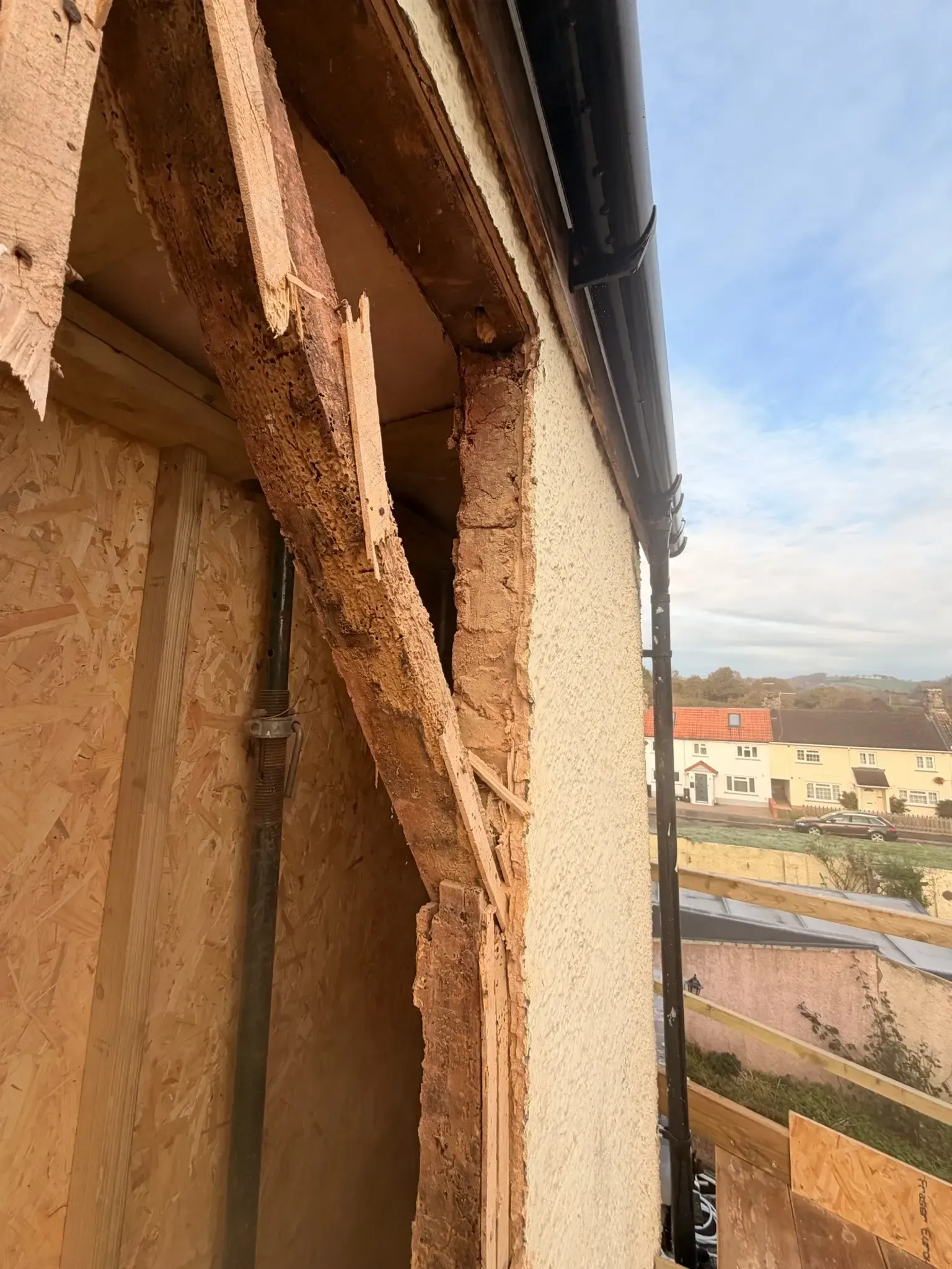 Damaged exterior wall with exposed insulation and wood framing, overlooking buildings under a blue sky