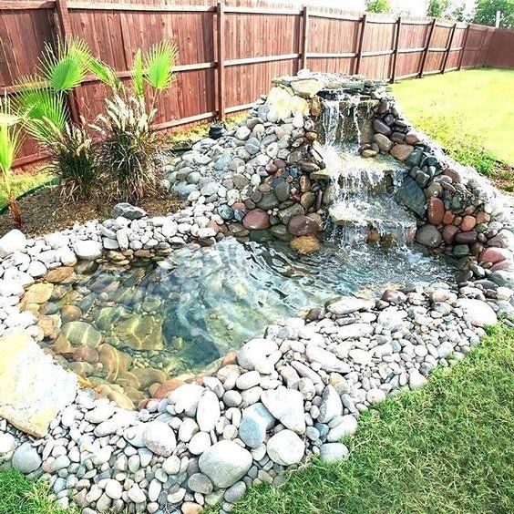 Rock-lined backyard pond waterfall with clear water, surrounded by grass and a wooden fence
