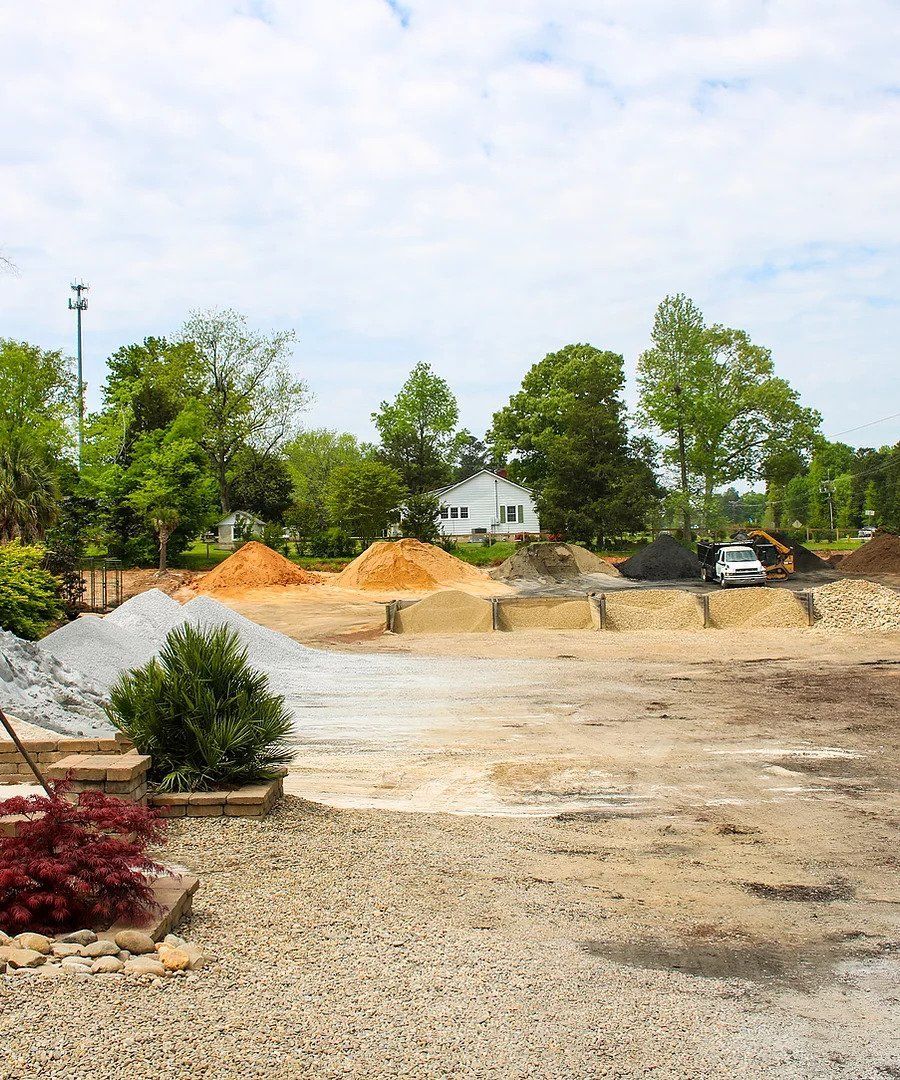 Gravel lot with piles of dirt, trees, and a small white house in the background under a cloudy sky