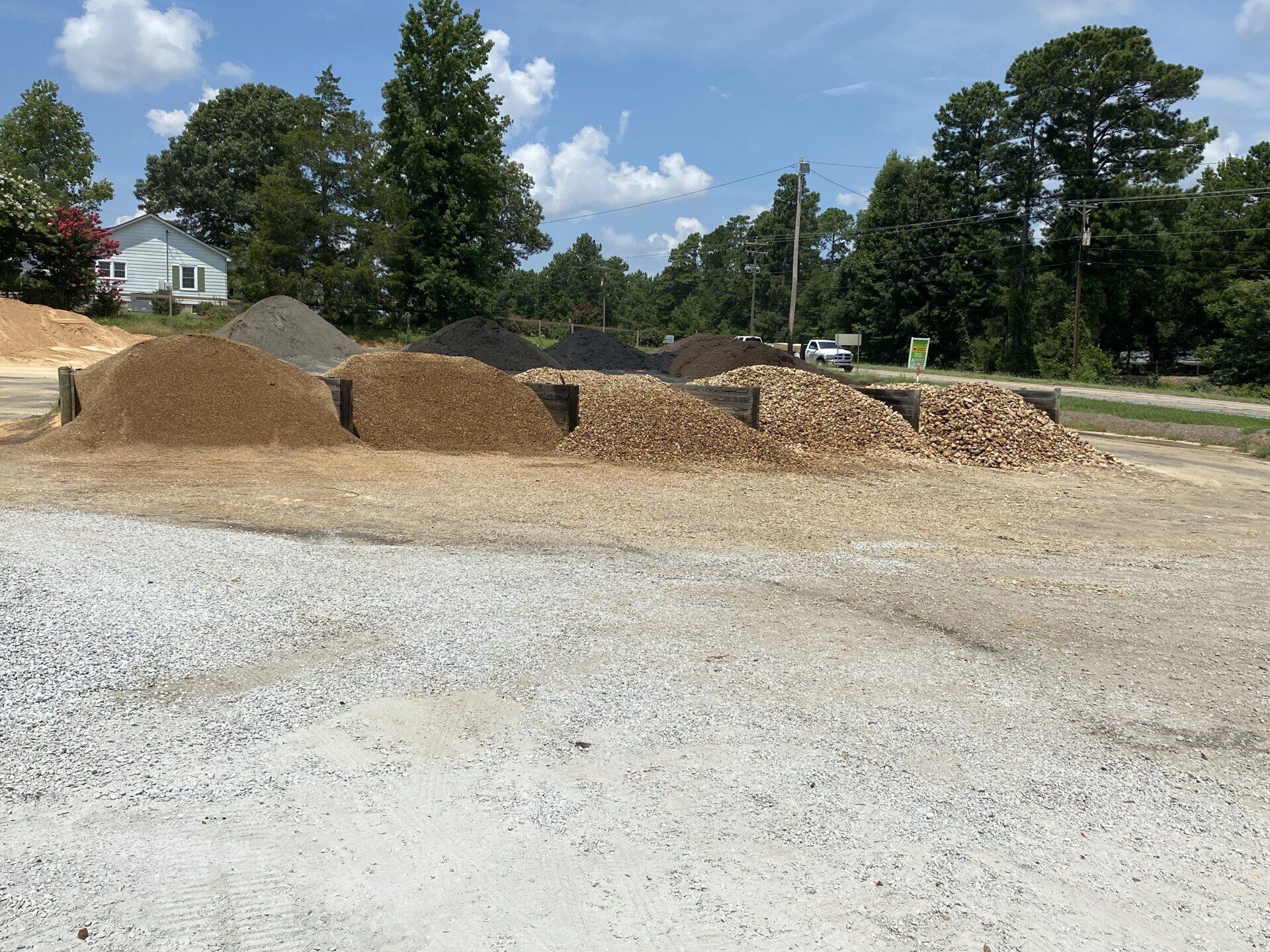 Pile of wood chips and gravel in a sunny outdoor lot with trees and houses in the background