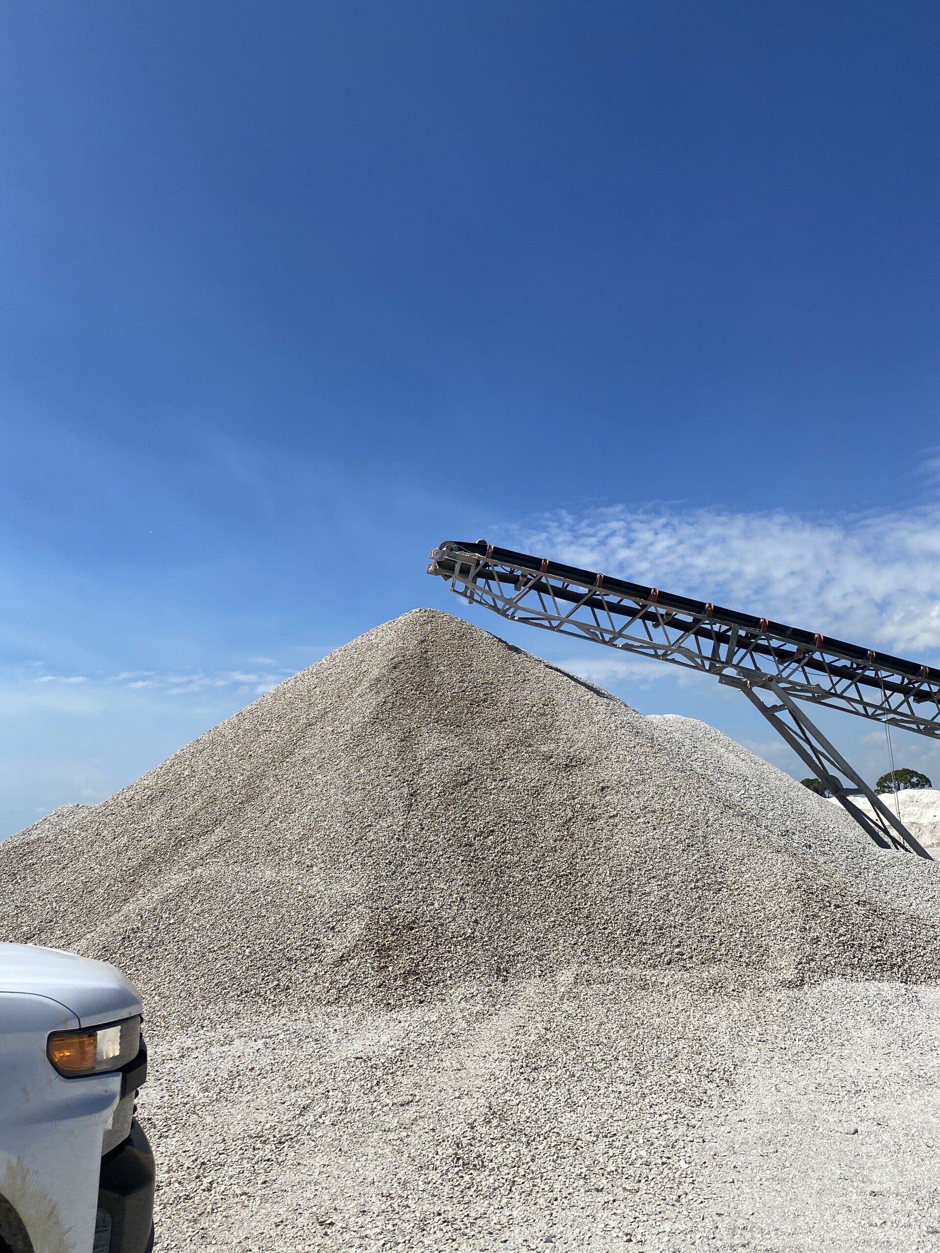 White gravel pile being loaded by a conveyor under a blue sky, with a truck at left.