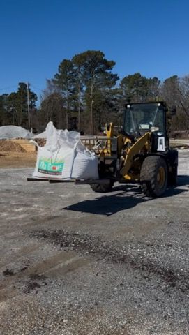 Yellow front loader lifting a large white bag on a gravel lot under a clear blue sky