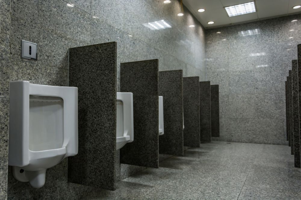 Una hilera de urinarios blancos en un limpio baño público gris con cubículos y paredes de azulejos moteados.
