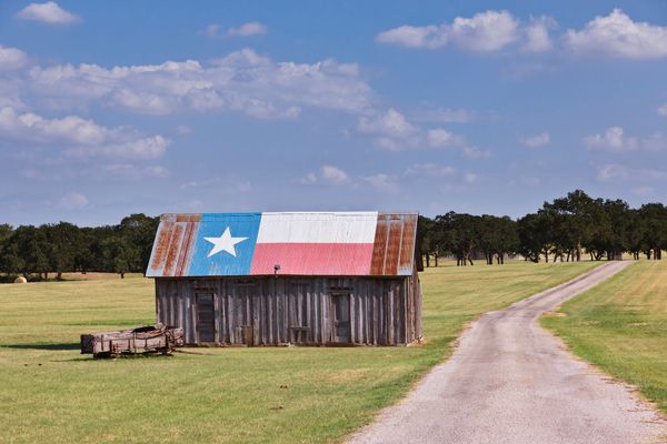 Barn with Texas flag painted on roof, dirt road, green field, and blue sky.