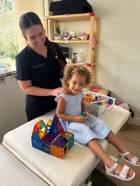 Child seated on a white treatment table with a clinician, holding a colorful toy in a bright therapy room