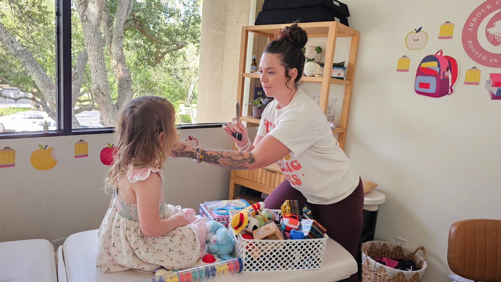 Adult handing a toy to a child in a bright playroom by a window.