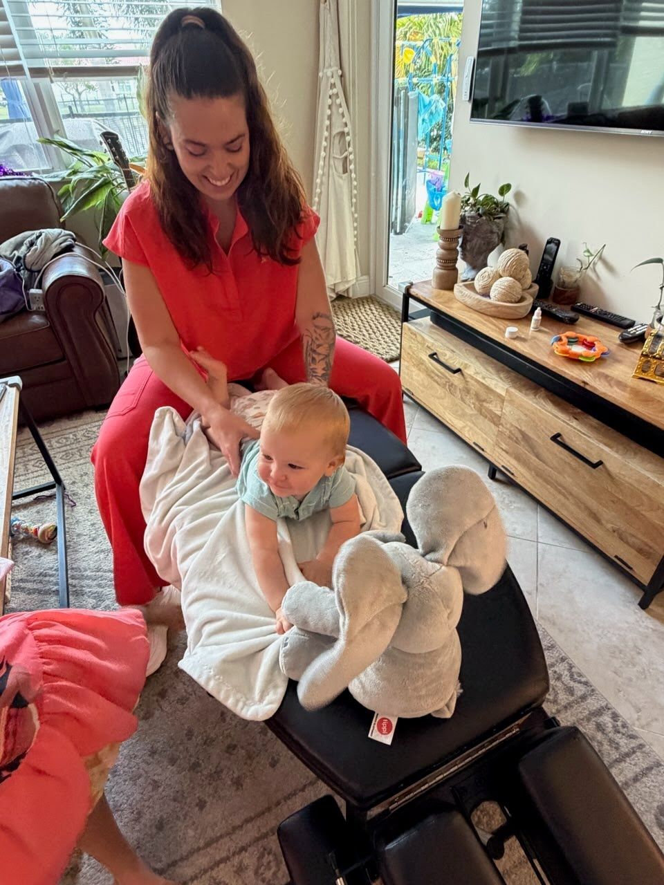 Woman in red helping baby sit on a black ottoman in a bright living room, with stuffed animals nearby