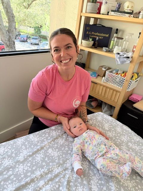 Smiling caregiver in pink shirt gently holds a baby on a bed in a bright room