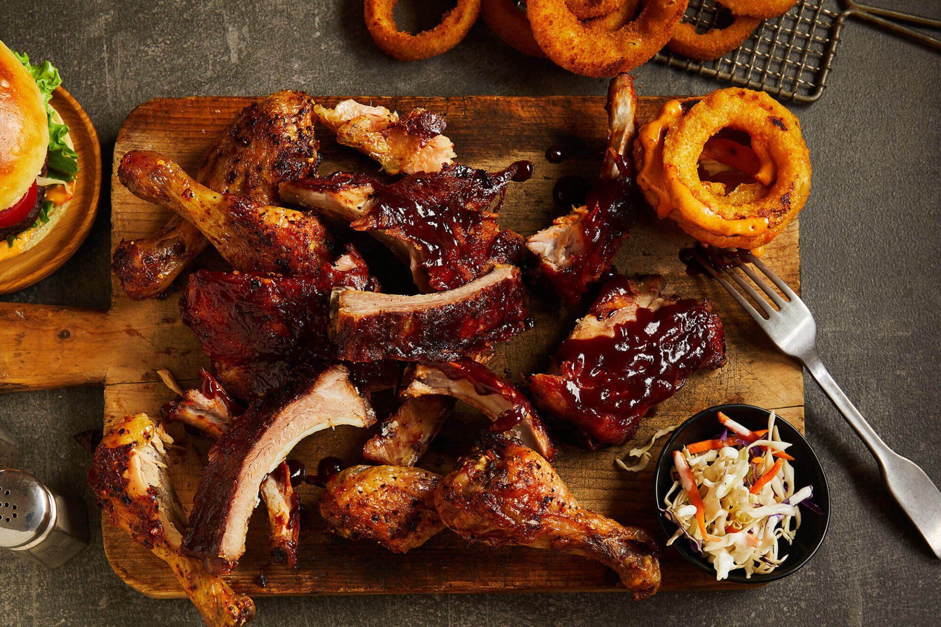 A wooden cutting board topped with ribs , chicken , onion rings and coleslaw.