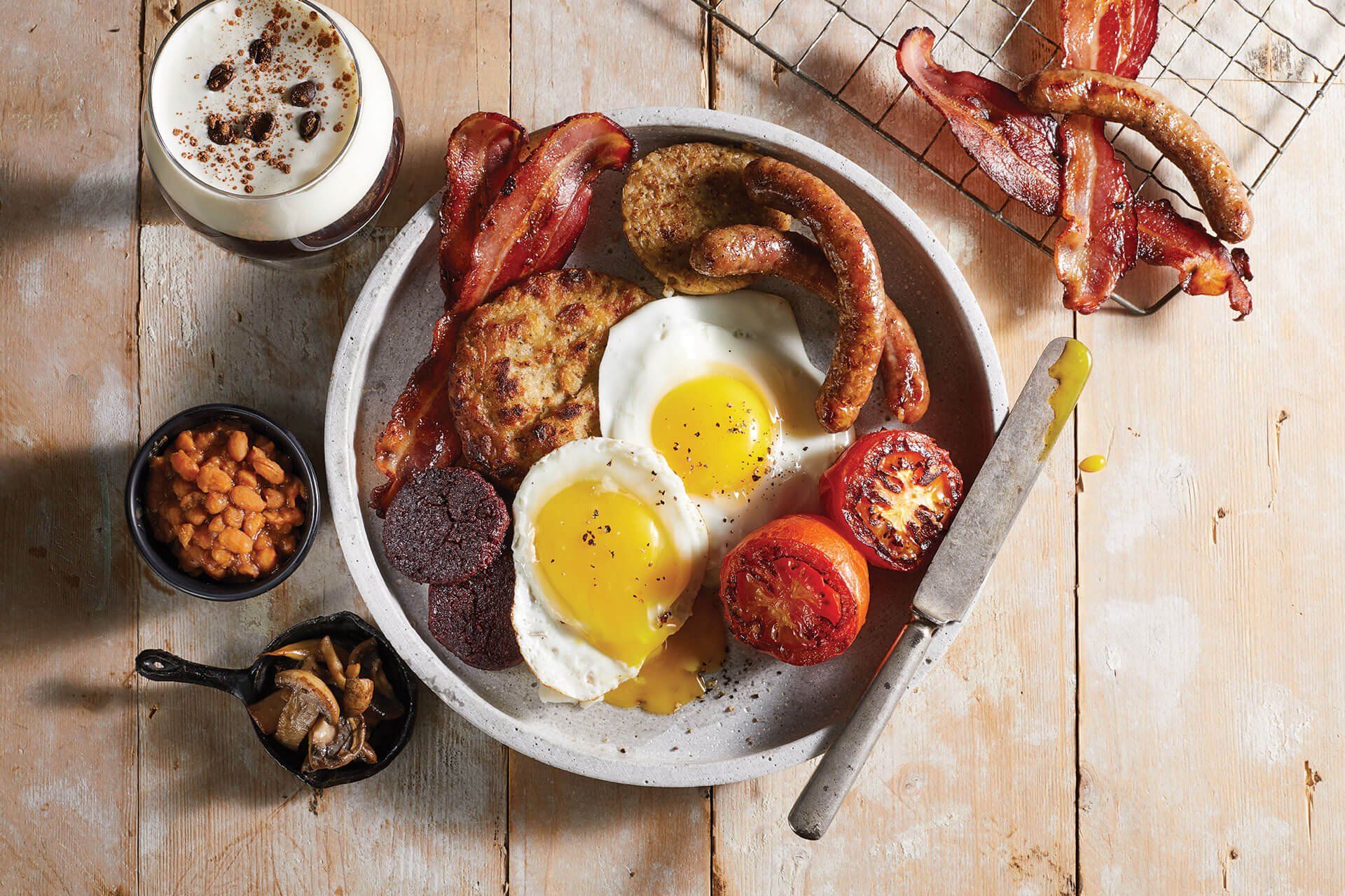 A plate of food with eggs , bacon , sausage , tomatoes and beans on a wooden table.
