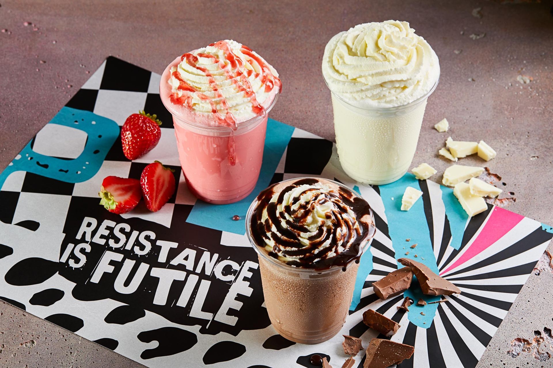 Three milkshakes are sitting on a table with strawberries and chocolate.