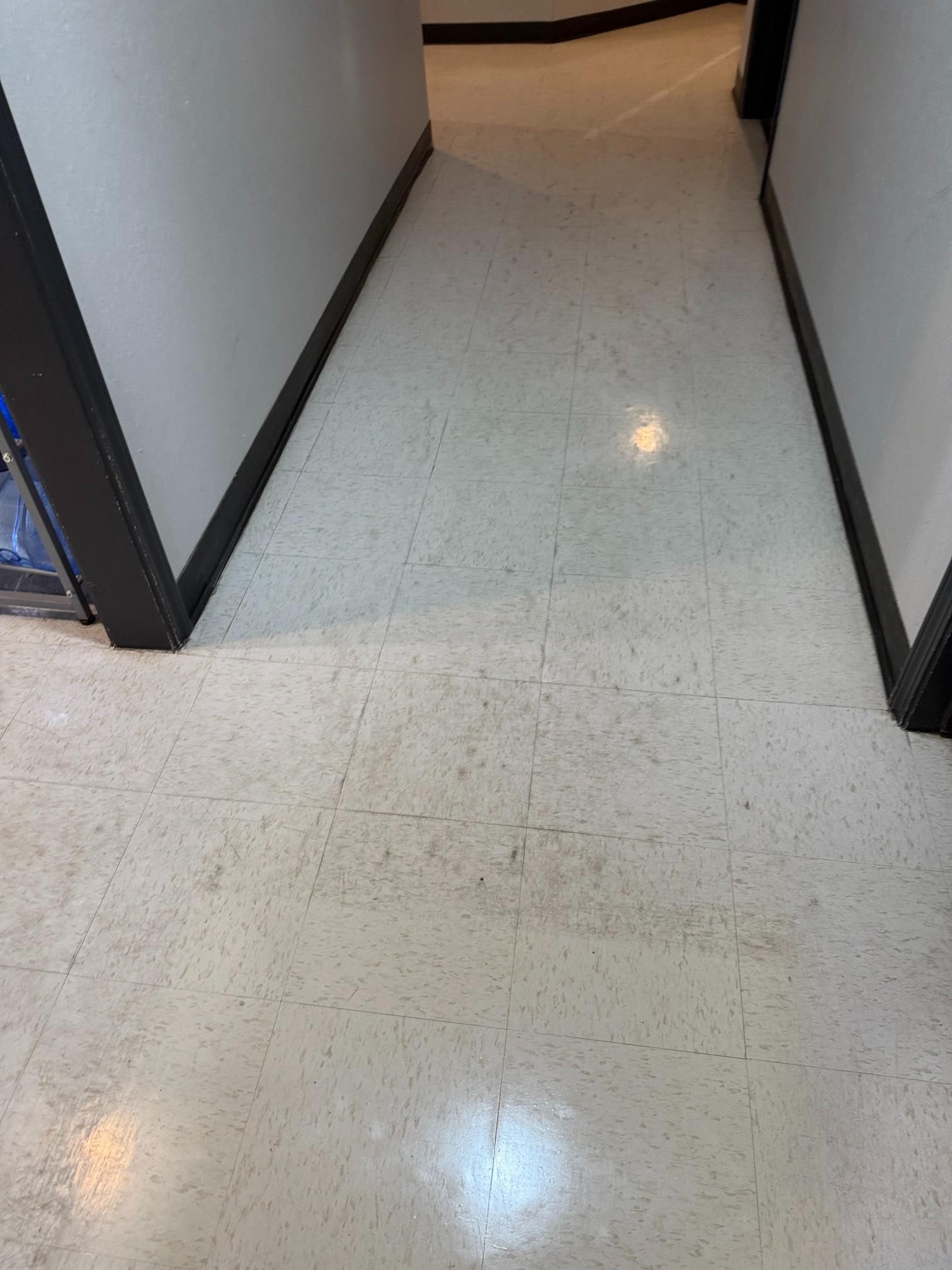 Narrow hallway with white speckled floor and gray walls, viewed toward a doorway.