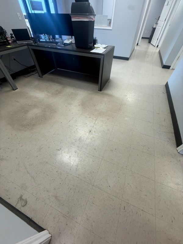 Empty office area with a black desk, computer monitors, and a large beverage dispenser on a beige floor.