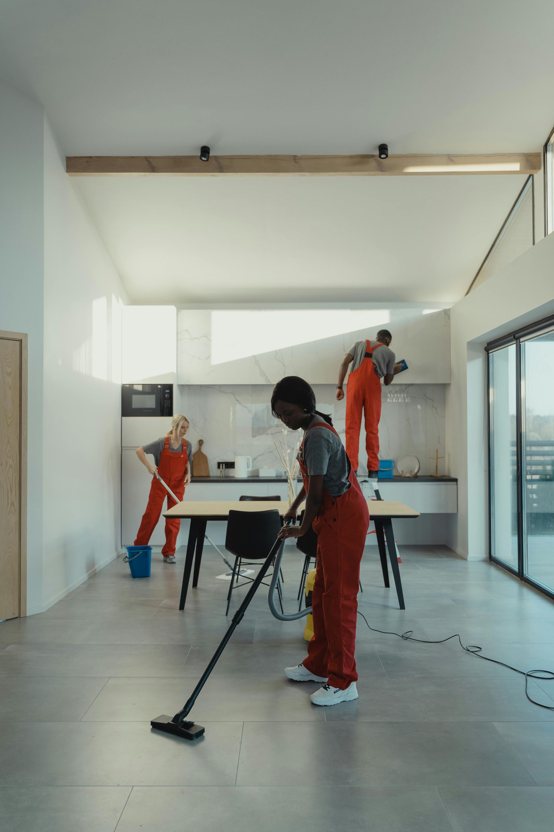 Three people in red jumpsuits cleaning a bright, modern kitchen. One vacuums, another mops, and the third wipes the cabinets.