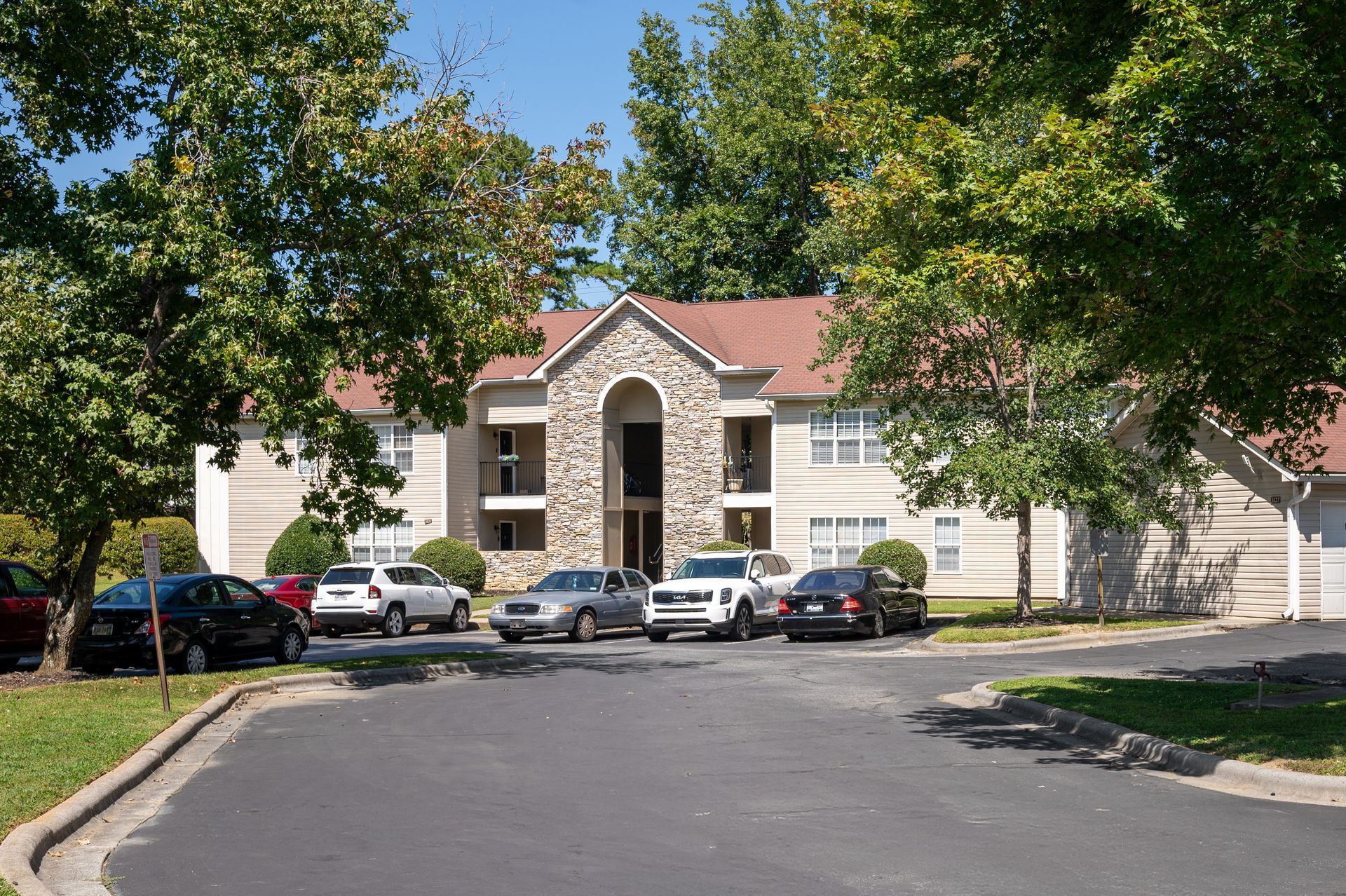 Stone apartment building with red roof and parked cars beneath leafy trees on a sunny day