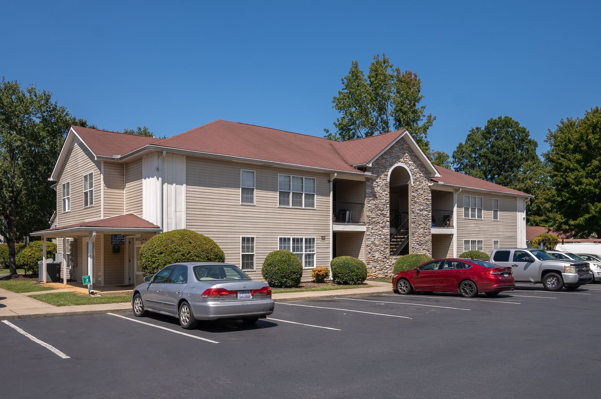 Apartment building with beige siding and red roof, parked cars in front on a sunny day