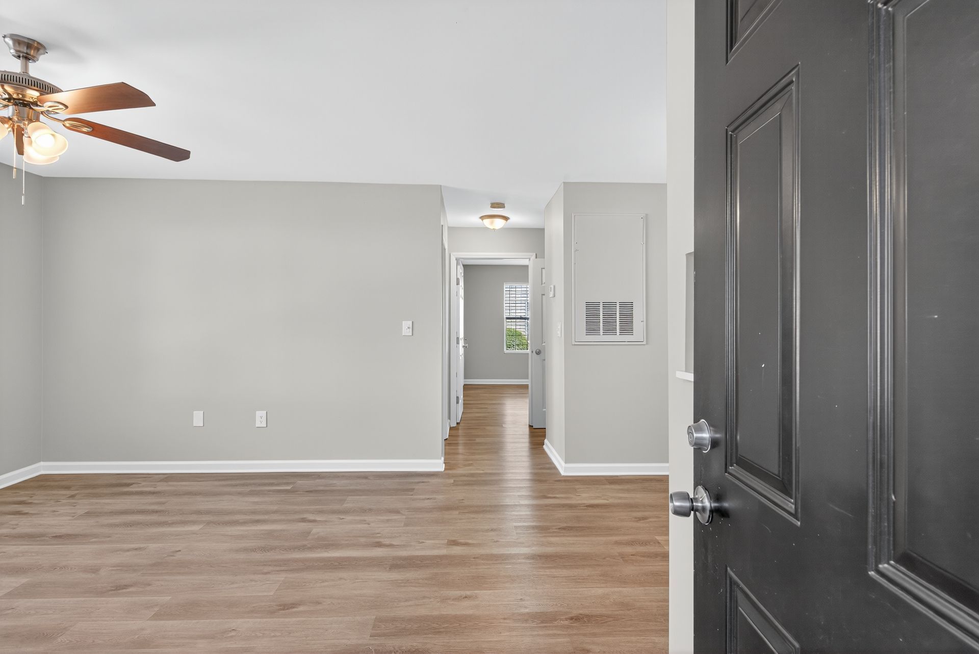 Empty living room with gray walls, wood flooring, ceiling fan, and open front door.